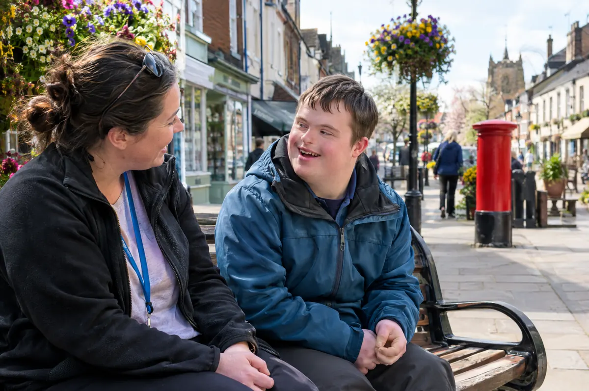 A young man and his support worker chatting on a bench on a sunny town high street